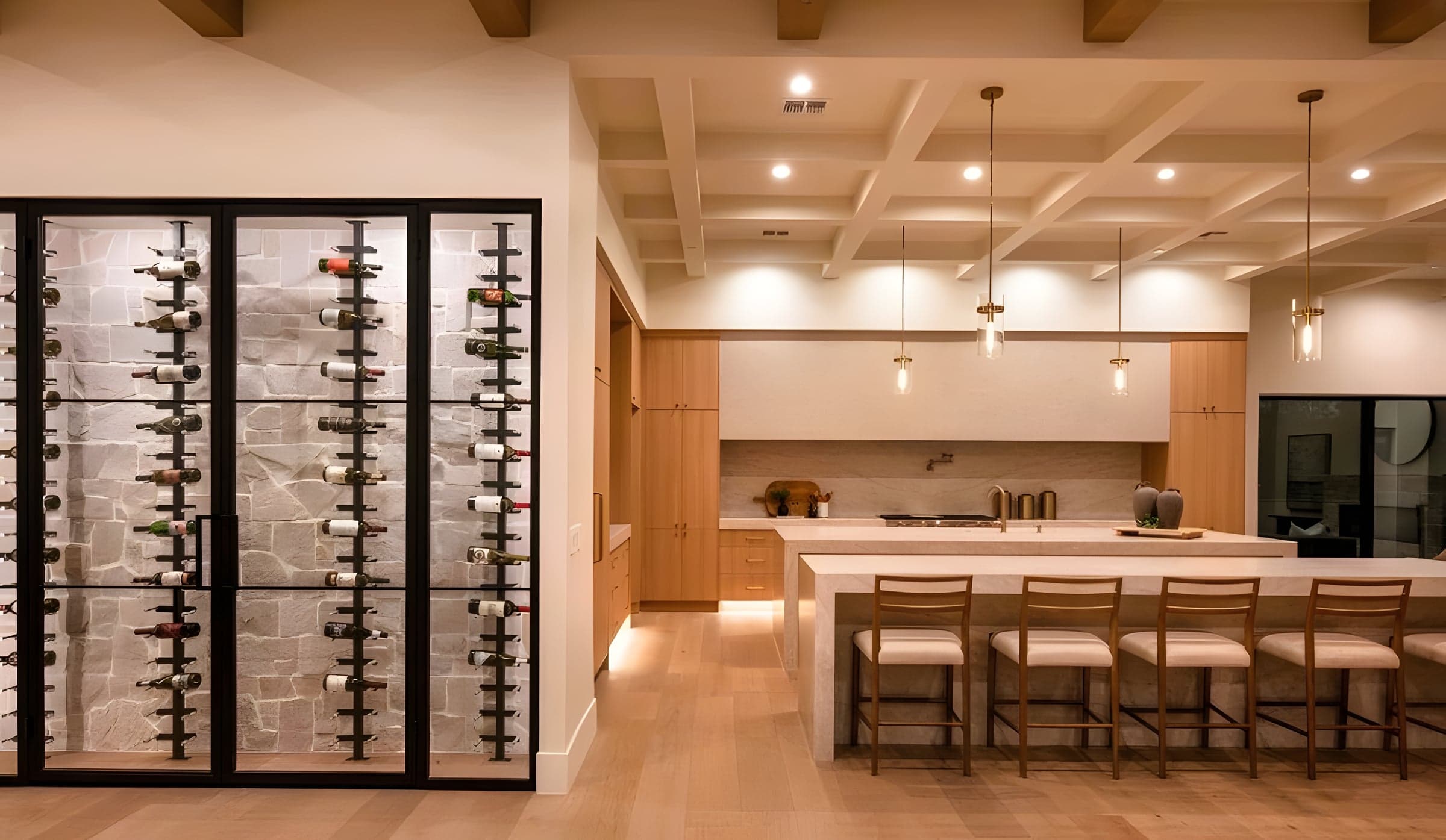The wine wall meets the kitchen at the entertaining heart of the home — stone, oak, brass, and pendant light in unhurried composition.