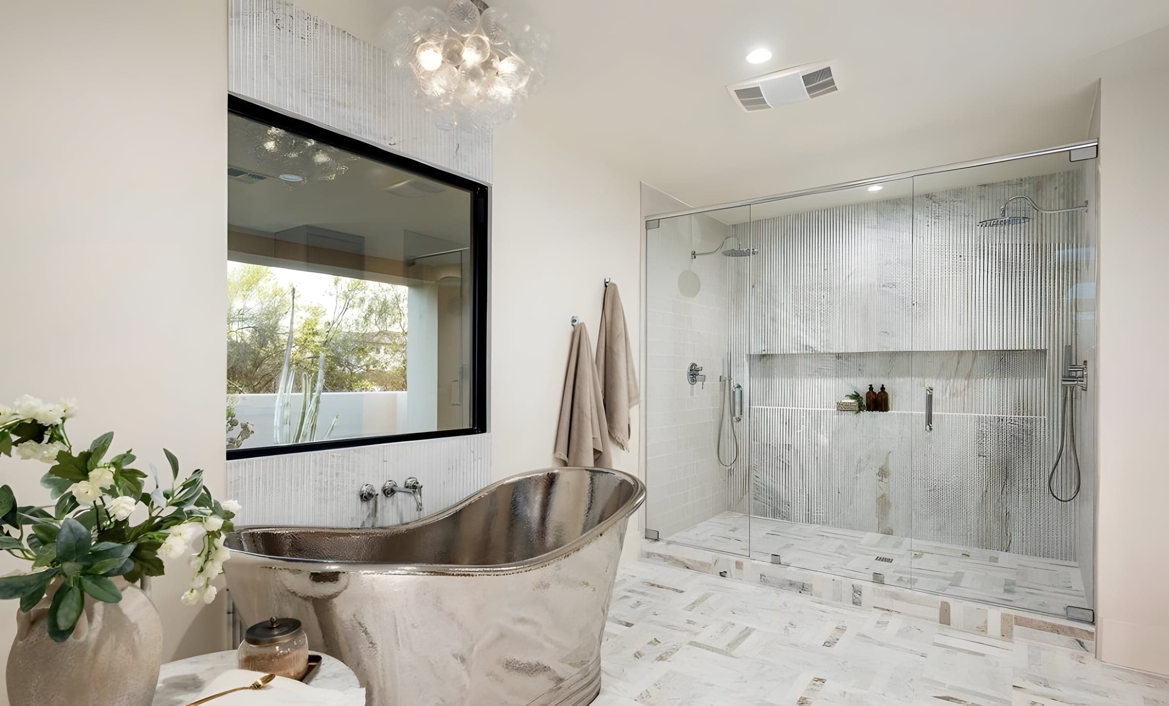 A hammered silver tub sits beneath a sculptural chandelier — fluted glass, herringbone marble, and brass fixtures complete the room.